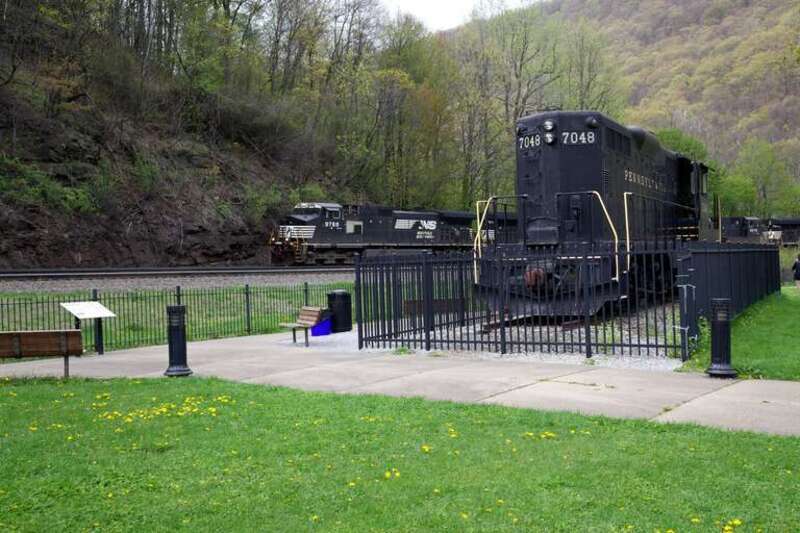 Monument of Pennsylvania RR locomotive 7048 - an EMD GP9 - with Norfolk Southern locomotive 9788 (GE Dash 9) leading a train around the curve.