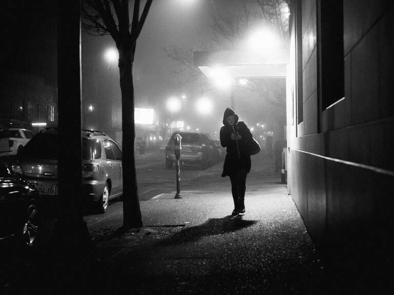 500px provided description: A woman walking down the street on a foggy night in the city. [#trees ,#city ,#street ,#night ,#lights ,#shadow ,#silhouette ,#woman ,#walking ,#refelction ,#streetlights ,#Fog ,#Coat]