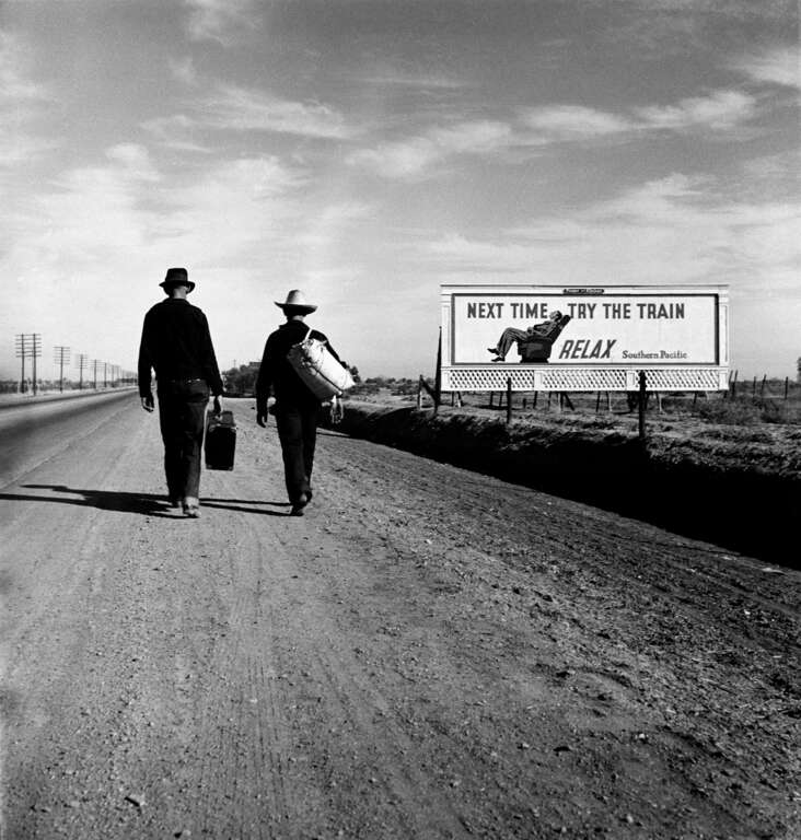 Photo shows two people walking along road near a billboard that says &quot;Next time, try the train. Relax.&quot; Nitrate negative. 2 1/4 x 2 1/4 inches.