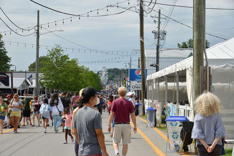 People walking down Maple Avenue, one of the main thoroughfares of the Montgomery County Agricultural Fair in Gaithersburg, Maryland.  on the right is &quot;The Big Cheese&quot; which sells grilled chees and block cheese, by the trees are the animal barns, and