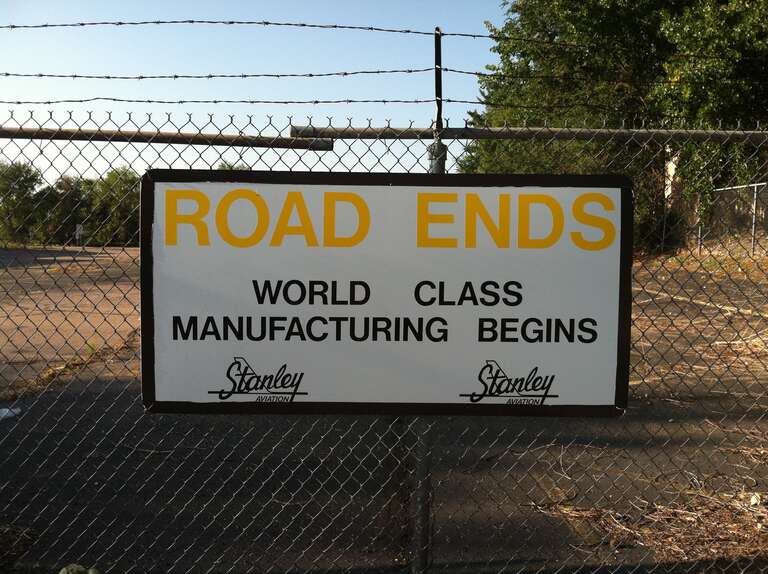 This is a sign attached to the chain link fence at the former Stanley Aviation plant at Stapleton Airport in Denver, Colorado.  The sign reads &quot;Road Ends World Class Manufacturing Begins&quot;.  The photo was taken August 25, 2012, long after the plant