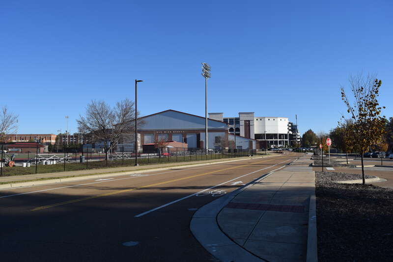 A road with sidewalks and bike lanes on the campus of the University of Mississippi adjacent to a parking lot and the Ole Miss Track and Field Facility. The Olivia and Archie Manning Athletics Performance Center and Vaught-Hemingway Stadium are in