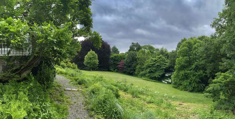 With a steep hill at its outer edge, a gravel-paved pathway clings to the south wall of the Glyndor Mansion, as seen on a June 2022 visit to the Wave Hill Public Garden. One of two historic homes located within this expertly manicured 28-acre