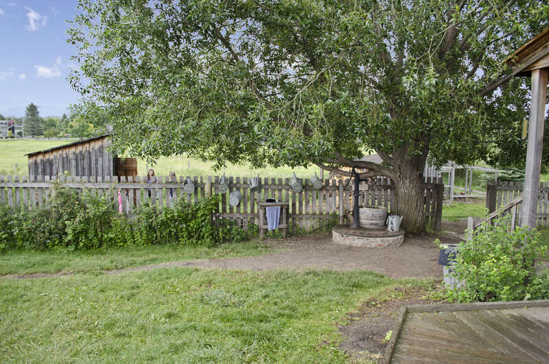 Looking southeast along the northeast side of the house on the grounds of the Tinsley Living Farm at the Museum of the Rockies in Bozeman, Montana.  The outhouse is the open-doored structure to the left.  The covered well is at the foot of the tree.
