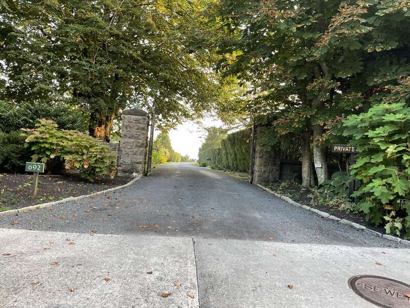 The photo depicts a residential driveway entrance during the daytime. The entrance is marked by a stone pillar on the left with the house number &quot;692&quot; displayed on a small green sign. On the right, another stone structure is partially obscured by