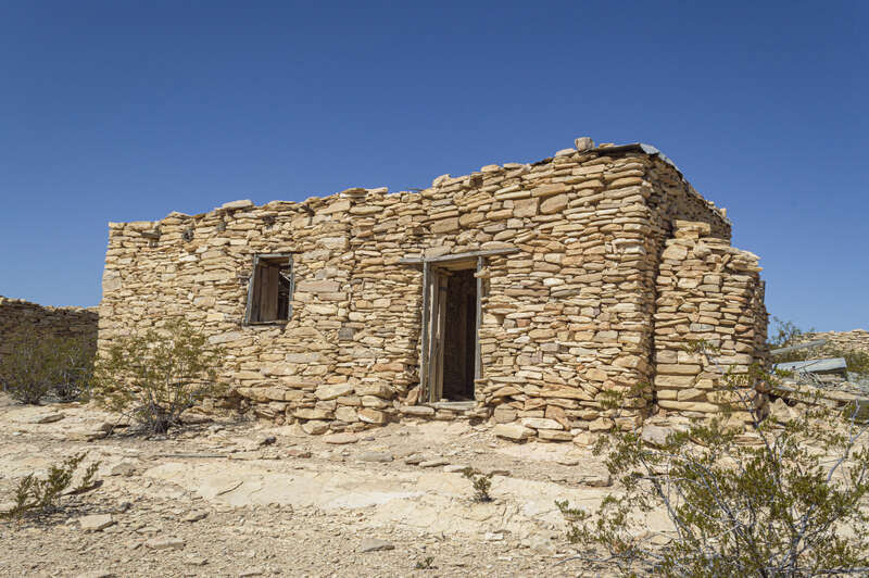A small 2 room house constructed of stacked limestone with a wooden post and lintel door and small window. The rock chimney seen on the right directs smoke from a fireplace.