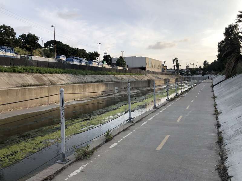 This is a picture of the Ballona Creek Bike Path in the evening during the summer. It was taken from the north bank, facing south. The concrete banks of the creek are clearly visible. Algae growth is visible in the creek. On the far bank, the rear