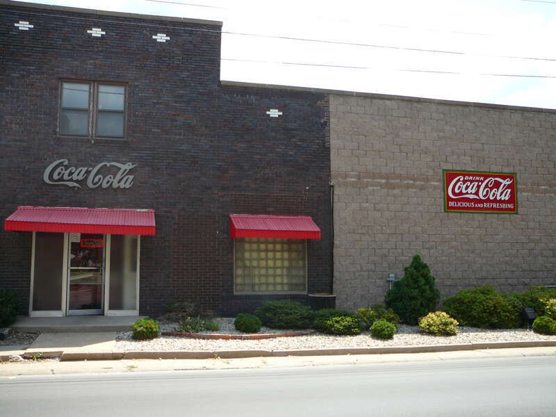 A photo of the front entrance and sign on adjacent building to the Coca-Cola Bottling Co of Macomb, a Coke distributor located in Macomb, IL 61455, taken from across the street on the west side of South Johnson