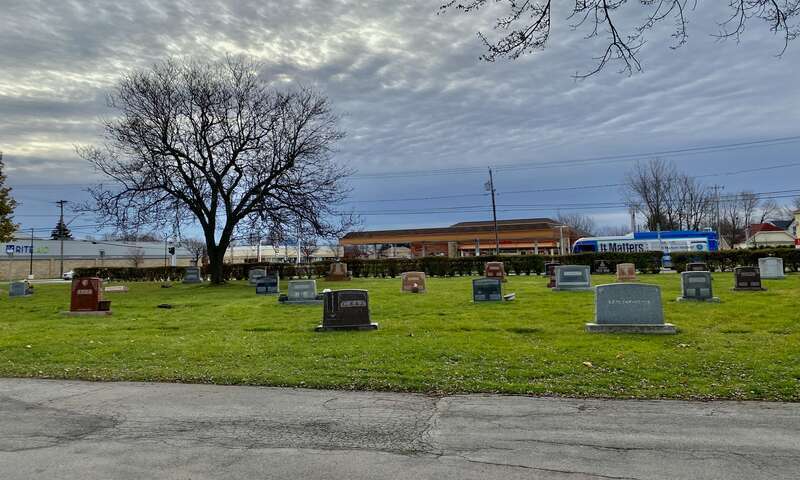 As seen from inside the Buffalo Cemetery in Cheektowaga, New York, an outbound NFTA Metro bus on route #24 nears the corner of Harlem Road on its eastward trajectory along Genesee Street in Cheektowaga, New York on a quiet Sunday morning in December