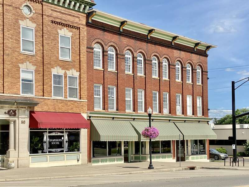 Built around the turn of the 20th Century, this Romanesque Revival and Italianate-style building features a red brick facade, bracketed cornice, low slope rear shed roof with a parapet, brick pilasters between the window bays, arched window bays not