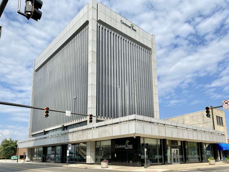Built in 1969, this Modern International-style skyscraper features a stone panel and metal-clad exterior with a one-story podium featuring a large parapet and dark stone-clad columns, a plaza atop the roof of the podium with planters and flagpoles,