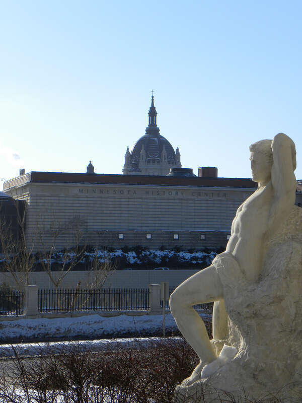 Minnesota History Center and the St. Paul Cathedral in the background with &quot;Earthbound, Made Captive, Yet Deserving Freedom More&quot; a 1956 marble sculpture by John Karl Daniels in the foreground on the Minnesota State Capitol Mall.