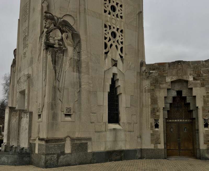 Looking diagonally at the base of the Charity Crucifixion Tower at the National Shrine of the Little Flower in Royal Oak, Michigan, showing examples of the Art Deco ornamentation on the exterior: from left to right, high-relief carvings of a Roman