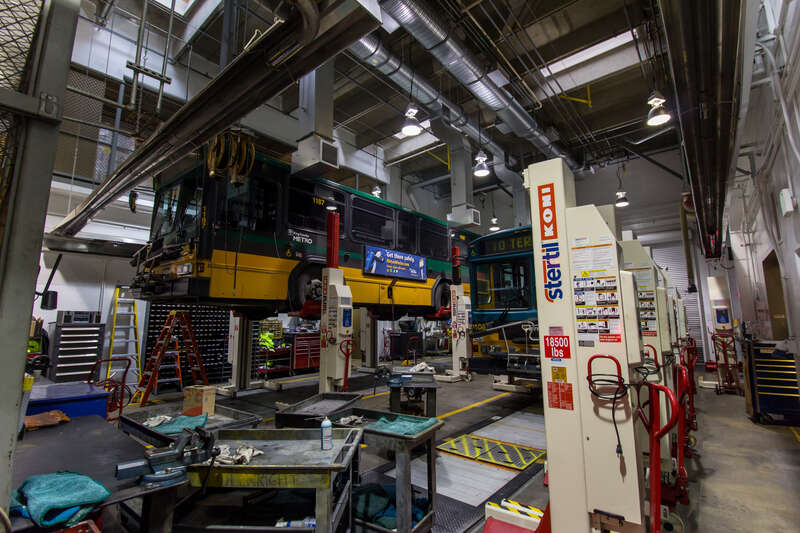 Two Gillig Phantom buses on vehicle lifts inside the Bellevue, Washington bus maintenance base for King County Metro, a transit provider in the Seattle area.