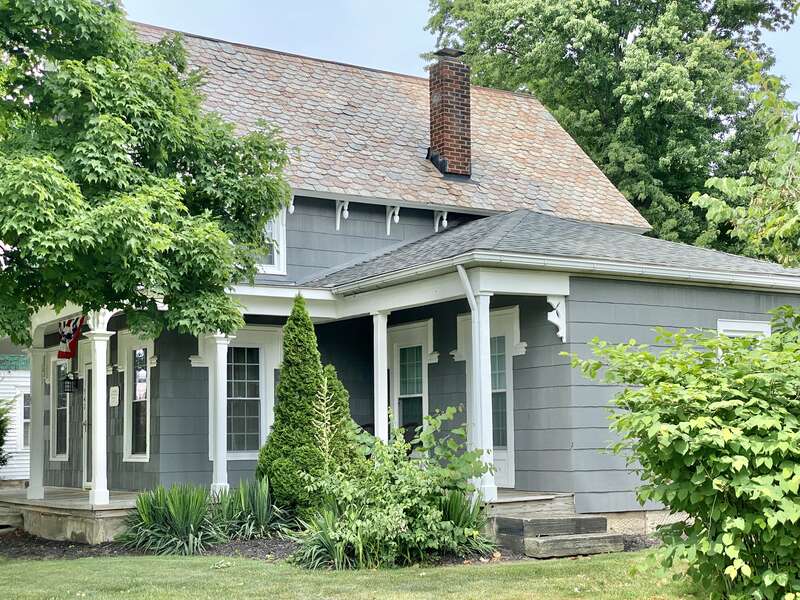 Built circa 1864, this Carpenter Gothic-style house features an exterior clad in asbestos shingles with ornate eave brackets, a front gable roof with decorative trim at the gable ends, decorative wooden window headers, a gothic arched window below